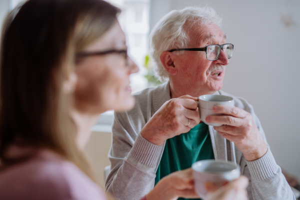 An adult daughter visiting her senior father at home and having coffee together, talking.