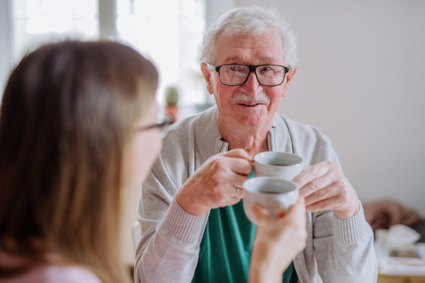 An adult daughter visiting her senior father at home and having coffee together, talking.