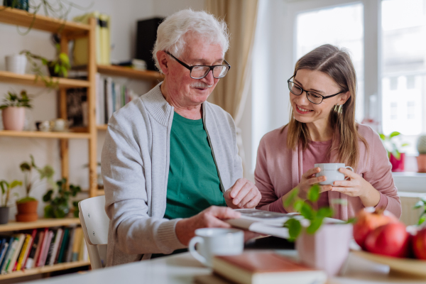 An adult daughter visiting her senior father at home and having coffee together, looking at book.