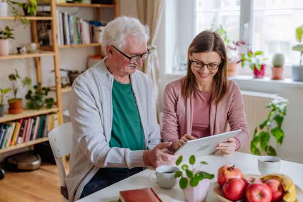 An adult daughter visiting her senior father at home and using tablet.