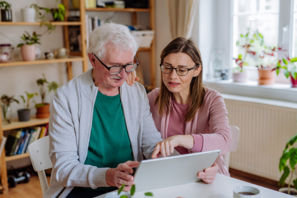 An adult daughter visiting her senior father at home and using tablet.