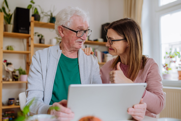 An adult daughter visiting her senior father at home and using tablet.