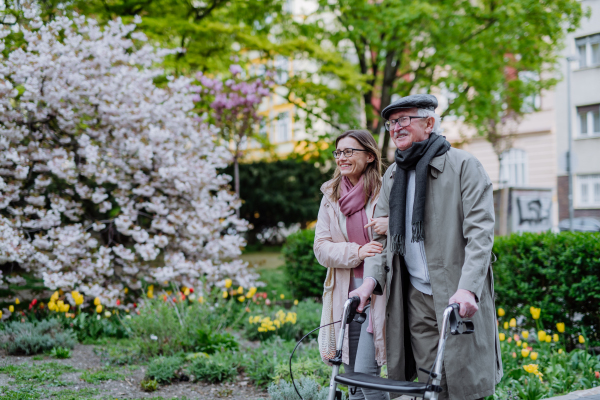 A senior man with walking frame and adult daughter outdoors on a walk in park.