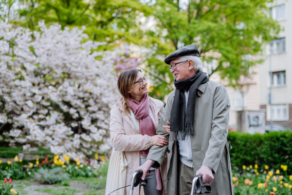 A senior man with walking frame and adult daughter outdoors on a walk in park.