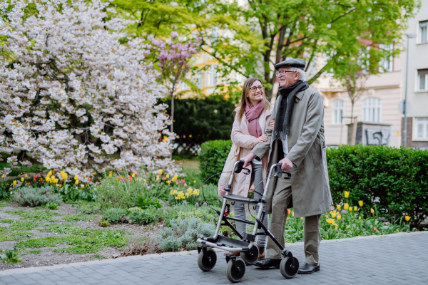 A senior man with walking frame and adult daughter outdoors on a walk in park.