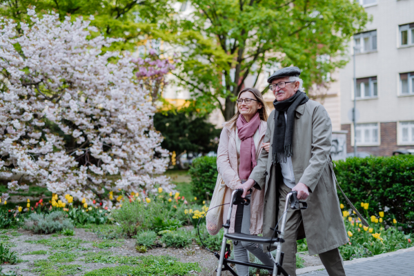 A senior man with walking frame and adult daughter outdoors on a walk in park.