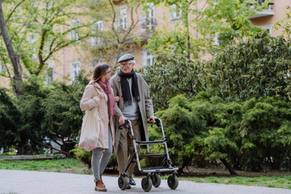 A senior man with walking frame and adult daughter outdoors on a walk in park.