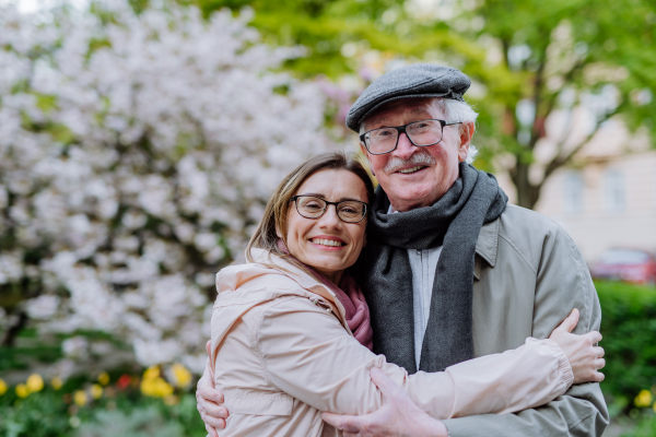 An adult daughter hugging her senior father outdoors in park on spring day.