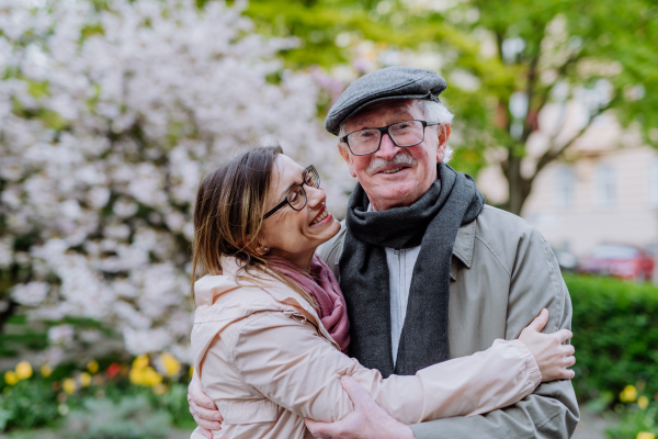 An adult daughter hugging her senior father outdoors in park on spring day.