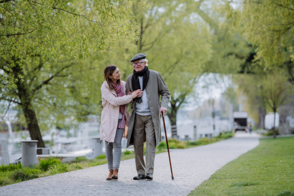 A happy senior man with walking stick and adult daughter outdoors on a walk in park.