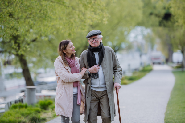 A happy senior man with walking stick and adult daughter outdoors on a walk in park.