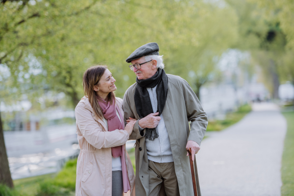 A happy senior man with walking stick and adult daughter outdoors on a walk in park.