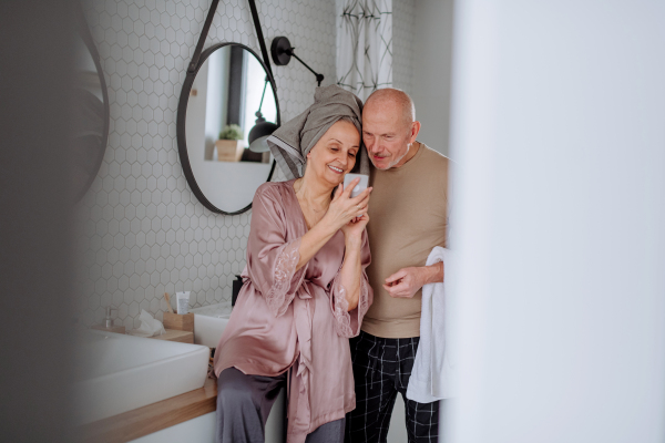 A senior couple in love in bathroom, using smartphone, morning routine concept.