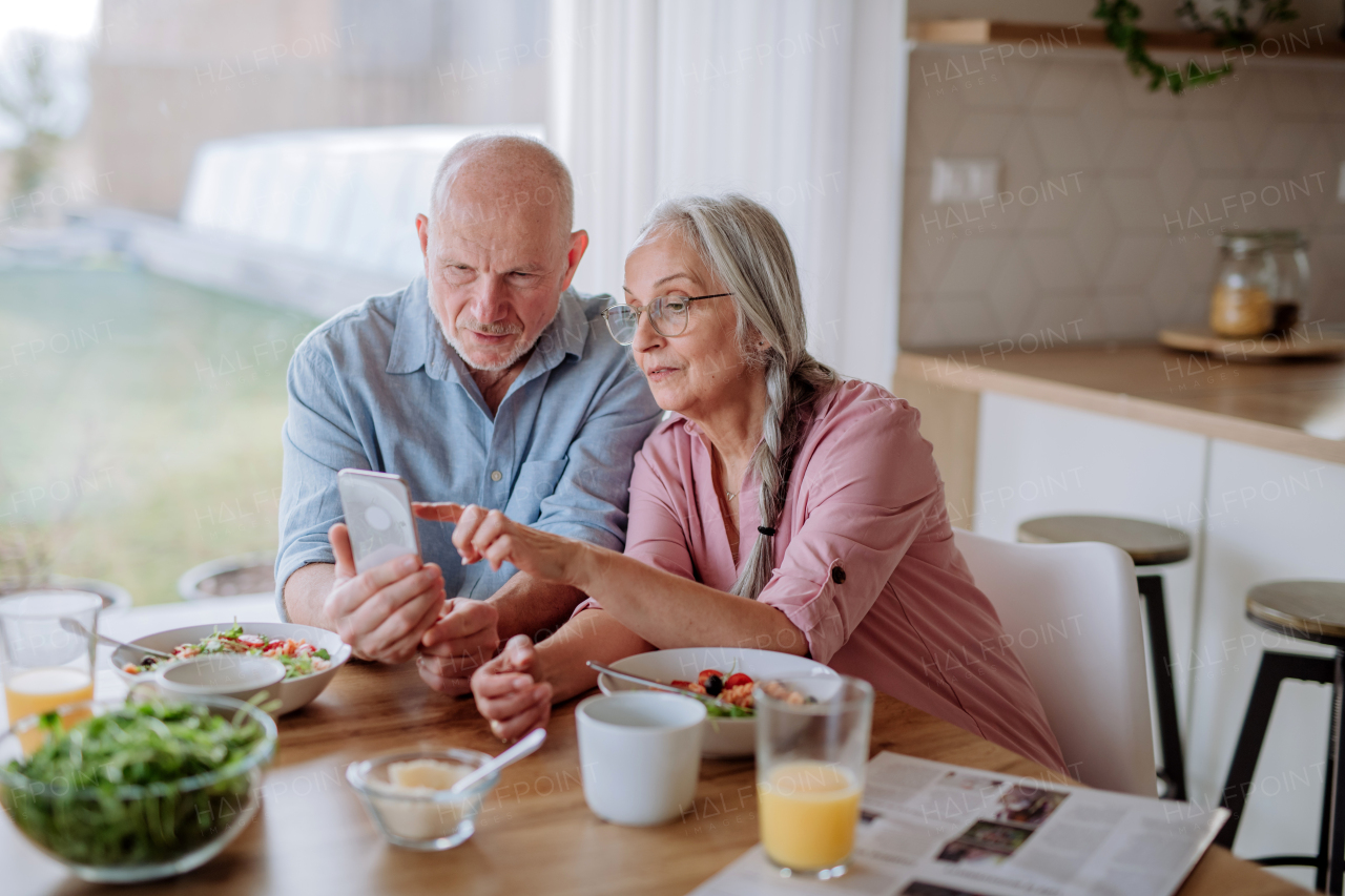 A happy senior couple using smartphone during eating dinner together at home.