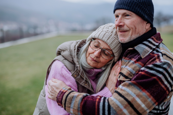 A senior man hugging and consoling his upset wife outdoors in garden in winter.