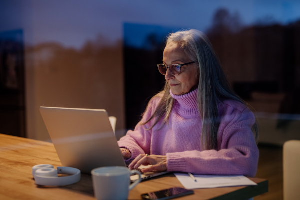 A senior woman sitting indoor and working on laptop.View trough the window.