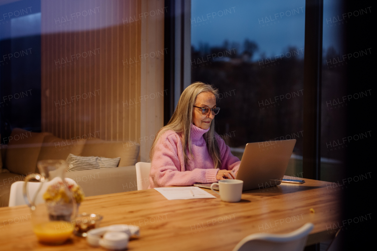 A senior woman sitting indoor and working on laptop.View trough the window.