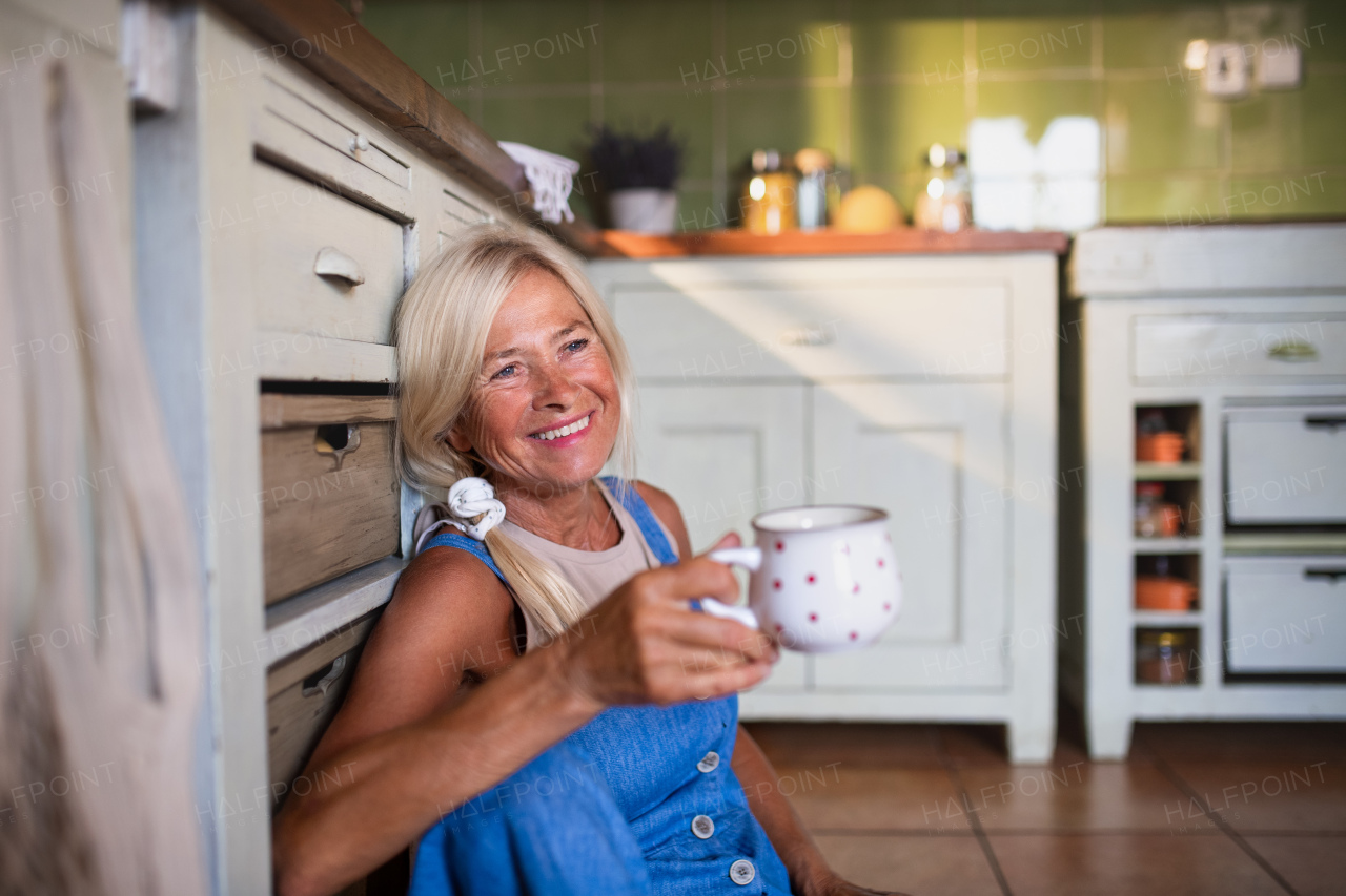 A happy senior woman sitting on floor and drinking tea indoors in kitchen.