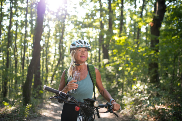 A happy senior woman biker drinking water from bottle outdoors in forest.