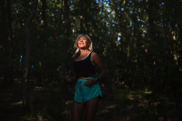 A happy active senior woman jogging outdoors in forest.