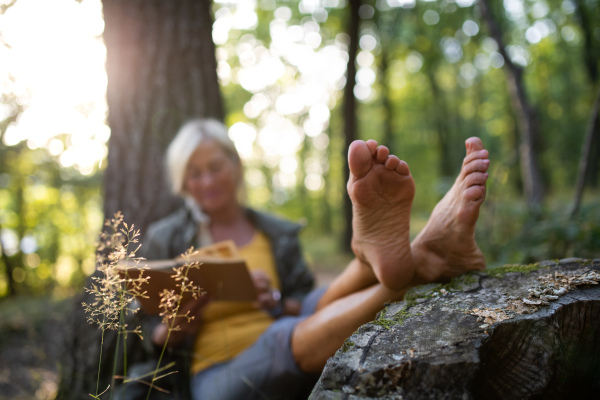 A senior woman relaxing and reading book outdoors in forest.