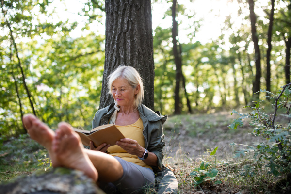 A senior woman relaxing and reading book outdoors in forest.