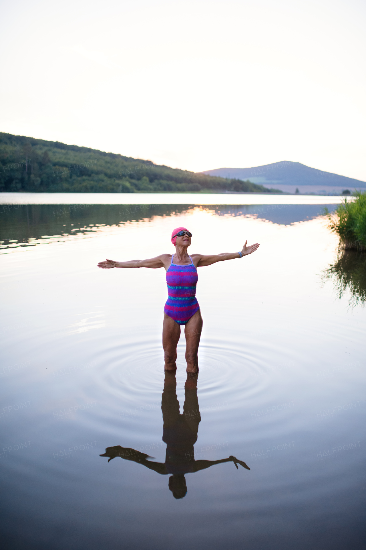 A portrait of active senior woman swimmer standing and stretching outdoors in lake.