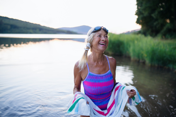 A portrait of active senior woman swimmer drying herself with towell outdoors by lake.