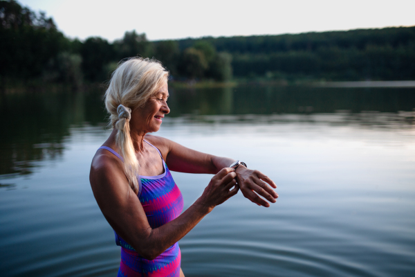 A portrait of active senior woman swimmer standing and setting smartwatch outdoors in lake.