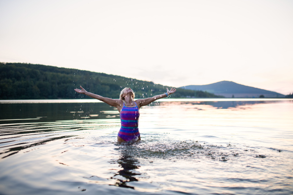 A portrait of active senior woman swimmer standing and stretching outdoors in lake.