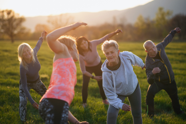 A group of seniors with sport instructor doing exercise outdoors in nature at sunset, active lifestyle.