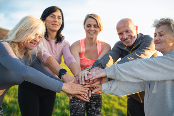 Group of seniors with sport instructor stacking hands together after exercise outdoors in nature, active lifestyle.