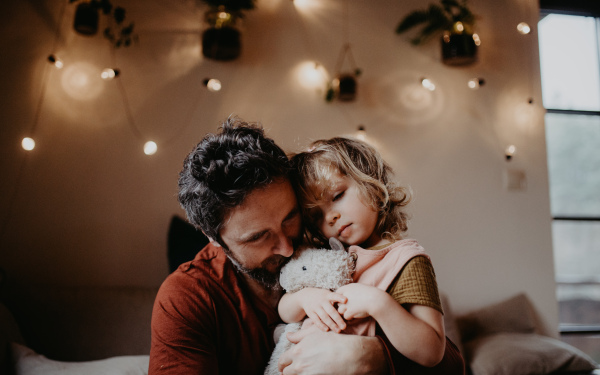 A mature father with sad small daughter hugging indoors at home.