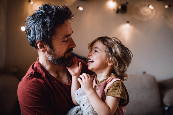 A mature father with small daughter standing indoors at home, holding and hugging.