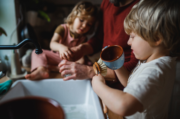 An unrecognizable father with two small children washing dishes indoors at home, daily chores concept.