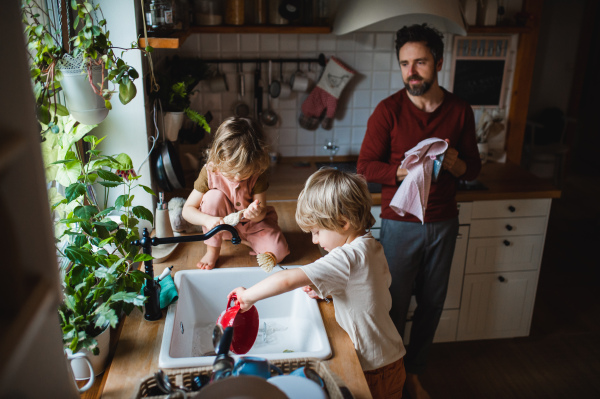 A mature father with two small children washing dishes indoors at home, daily chores concept.