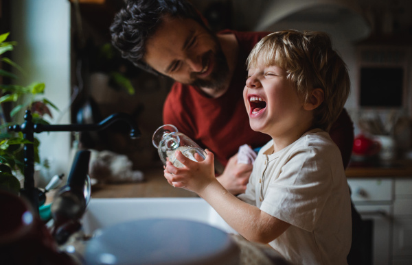 A small laughing boy with father having fun when washing dishes indoors at home, daily chores concept.