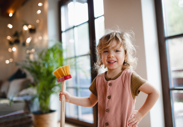 A small girl with broom helping with housework indoors at home, looking at camera.