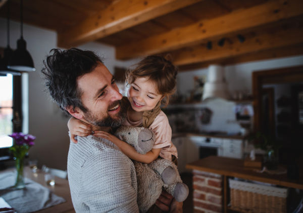 A mature father with small daughter standing indoors at home, holding and hugging.