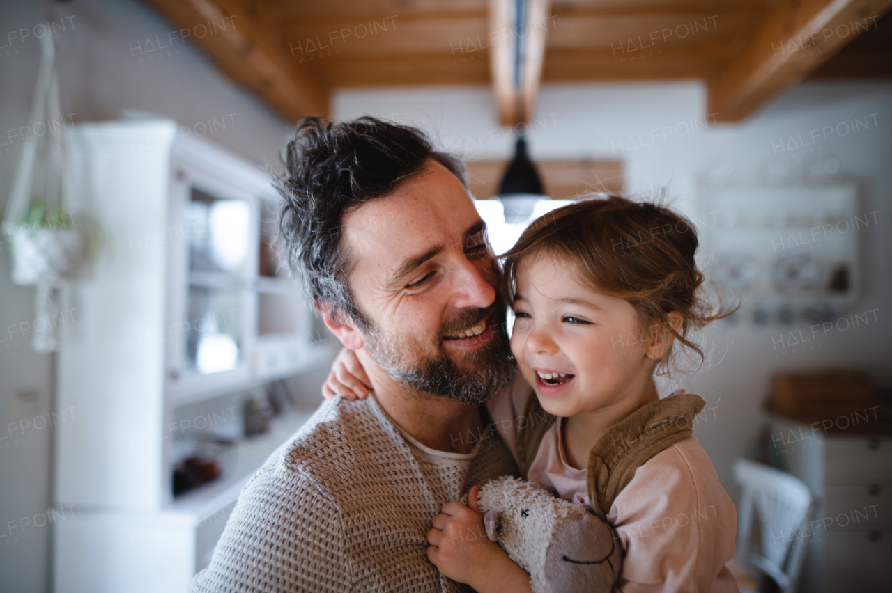 A mature father with small daughter standing indoors at home, holding and hugging.