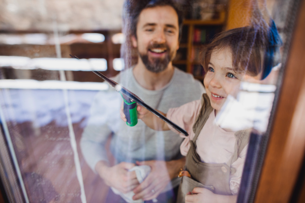 A happy girl with father cleaning windows at home, daily chores concept. Shot through glass.