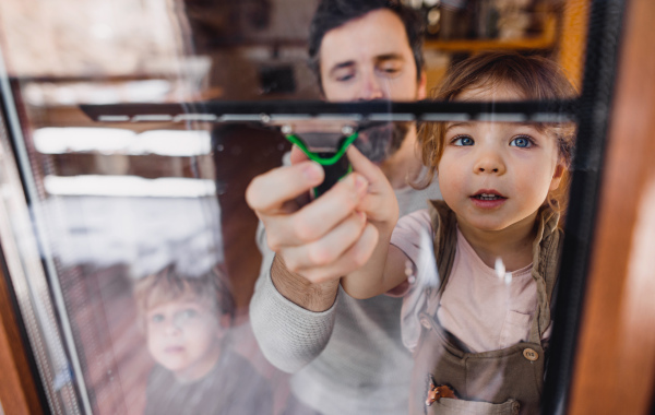 A happy girl with father cleaning windows at home, daily chores concept. Shot through glass.