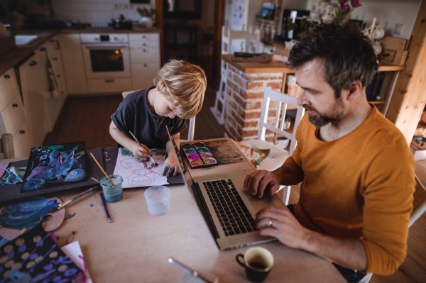 Mid adult father with a small son working indoors at home, painting pictures and home office concept.