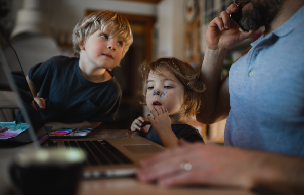 An unrecognizable father with two small children working indoors at home, painting pictures and home office concept.