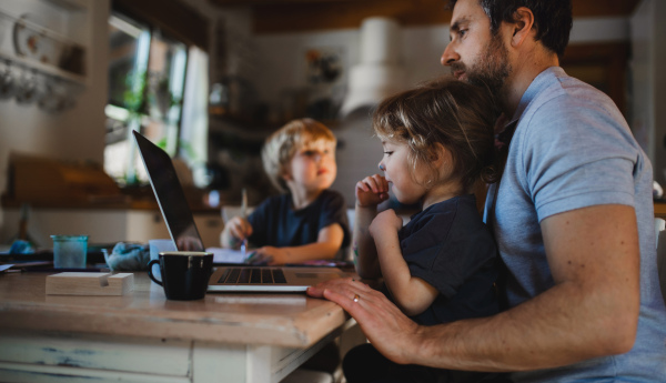 A side view of mid adult father with small children working indoors at home, home office concept.