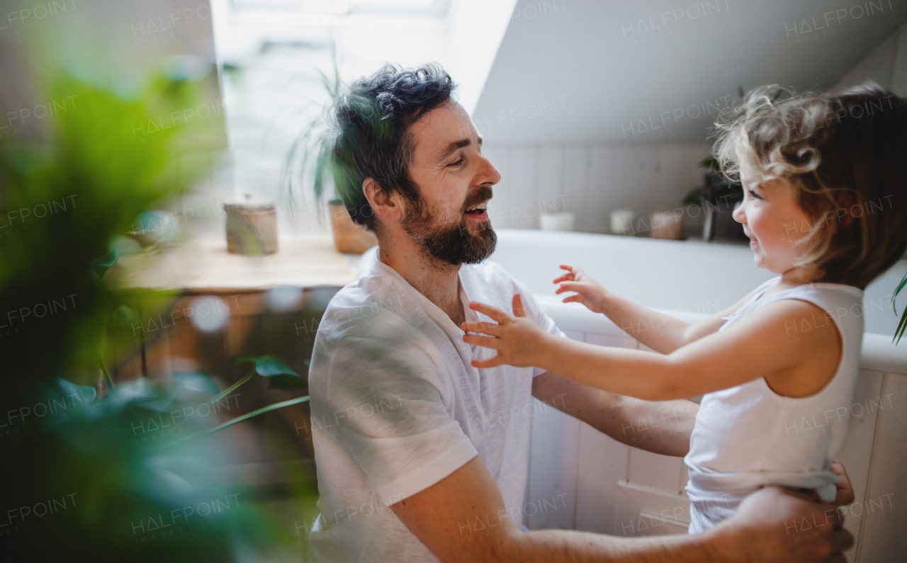 Side view of mature father with small daughter indoors at home, getting ready for a bath.