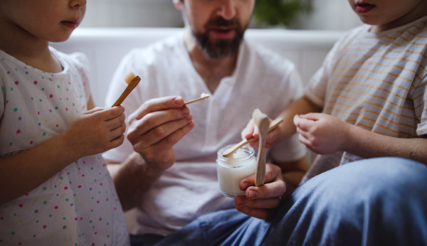 An unrecognizable father with two small children brushing teeth indoors at home, sustainable lifestyle concept.