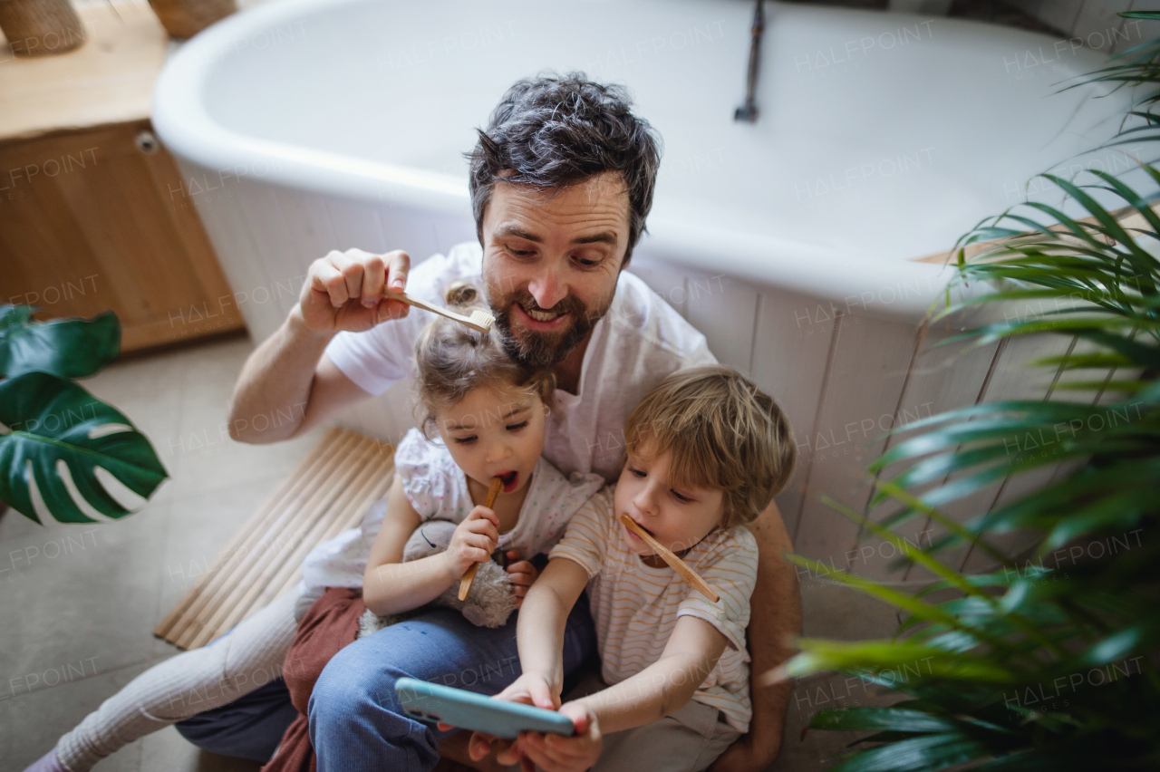 High angle view of a father with two small children brushing teeth indoors at home and taking selfie, sustainable lifestyle concept.