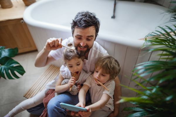 High angle view of a father with two small children brushing teeth indoors at home and taking selfie, sustainable lifestyle concept.