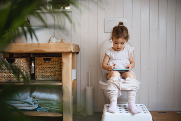 A portrait of cute small sitting on toilet indoors at home, using smartphone.